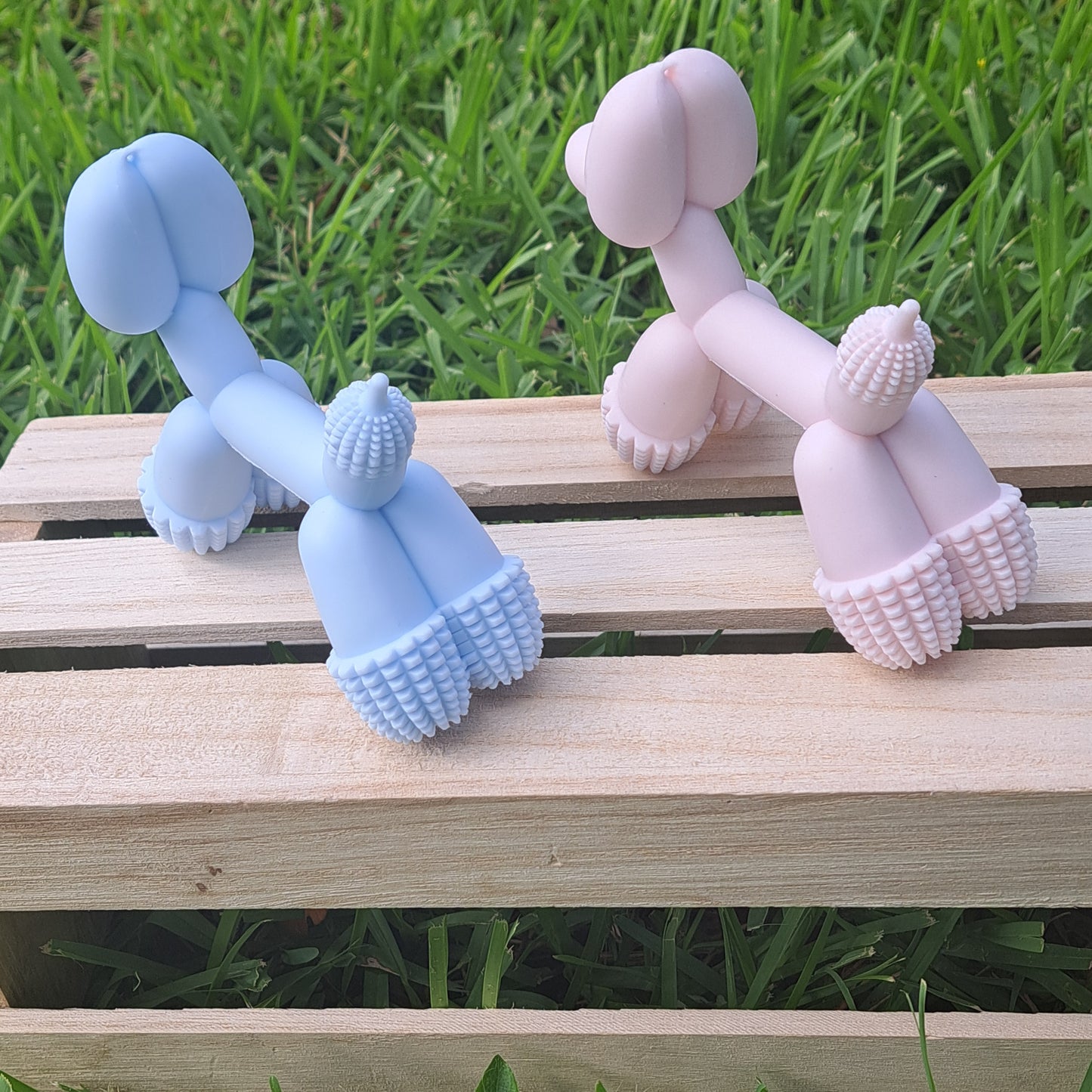 Two silicone dog toys, one pink and one blue, on a wooden surface with grass in the background.