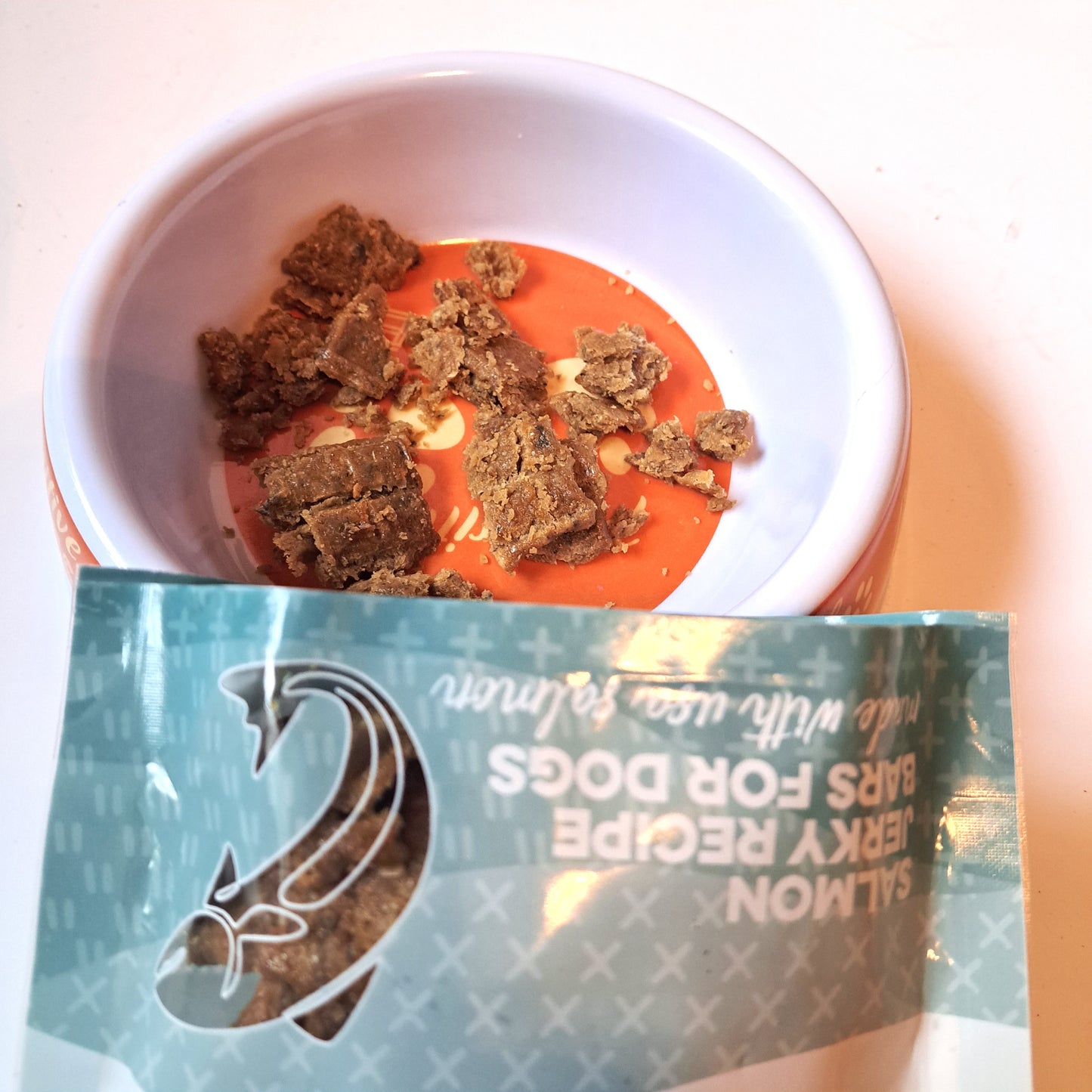 A bowl of dog jerky treats with wild-caught salmon flavor next to a packaging bag with the product details visible.