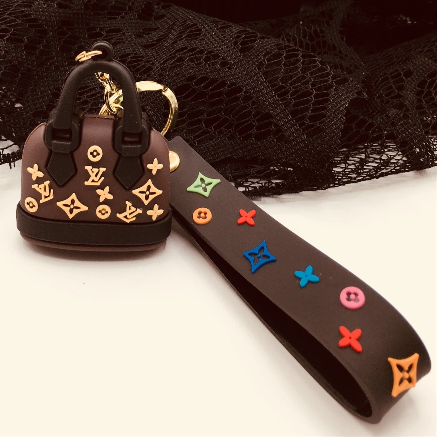 A small designer handbag keychain charm in the shape of a handbag, with gold-colored hardware, placed on a black fabric surface next to a pumpkin and a decorative tree.