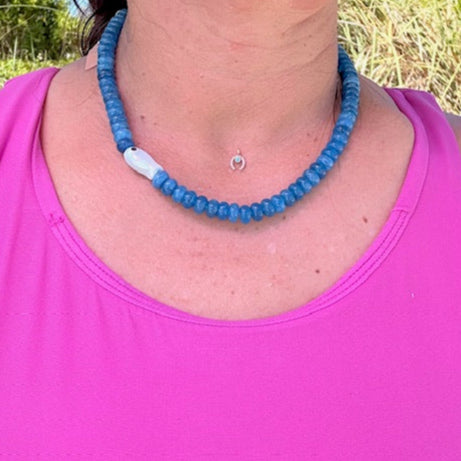 Person wearing a blue beaded necklace outdoors with greenery in the background