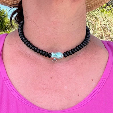Person wearing a black beaded necklace with a turquoise charm outdoors