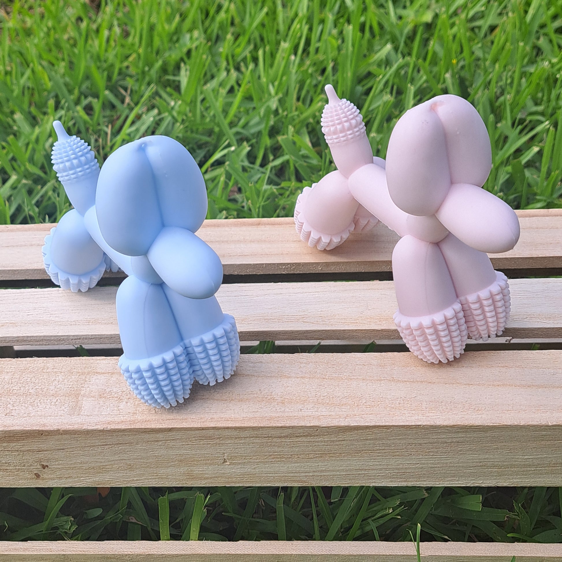 Two dog toy figures, one pink and one blue, on a wooden surface with grass in the background.
