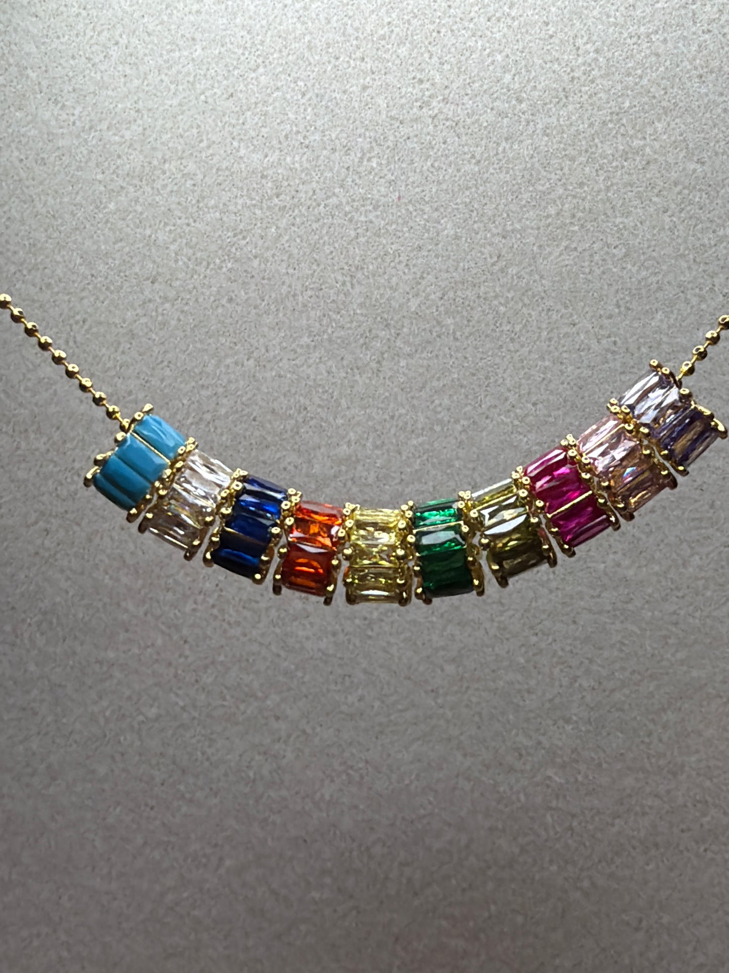 A gold chain necklace featuring a row of colorful baguette-cut gemstones in blue, clear, orange, yellow, green, olive, and pink, displayed against a textured gray background.