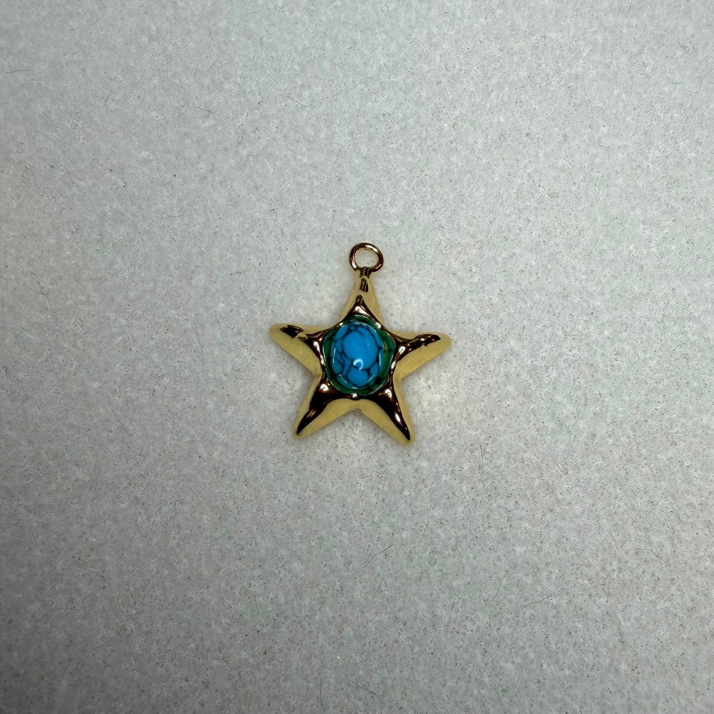 A small gold star-shaped charm pendant with a central blue oval gemstone, resting on a textured light gray surface.